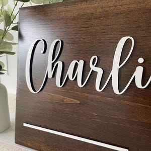 May include: A dark brown wooden sign with the name "Charli" in white, cursive lettering. The sign has a white horizontal line at the bottom. The wood grain is visible. The sign is leaning against a white surface.