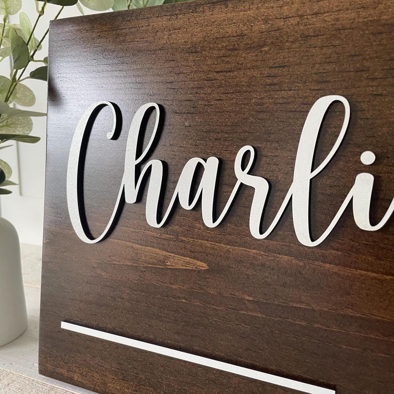 May include: A dark brown wooden sign with the name "Charli" in white, cursive lettering. The sign has a white horizontal line at the bottom. The wood grain is visible. The sign is leaning against a white surface.