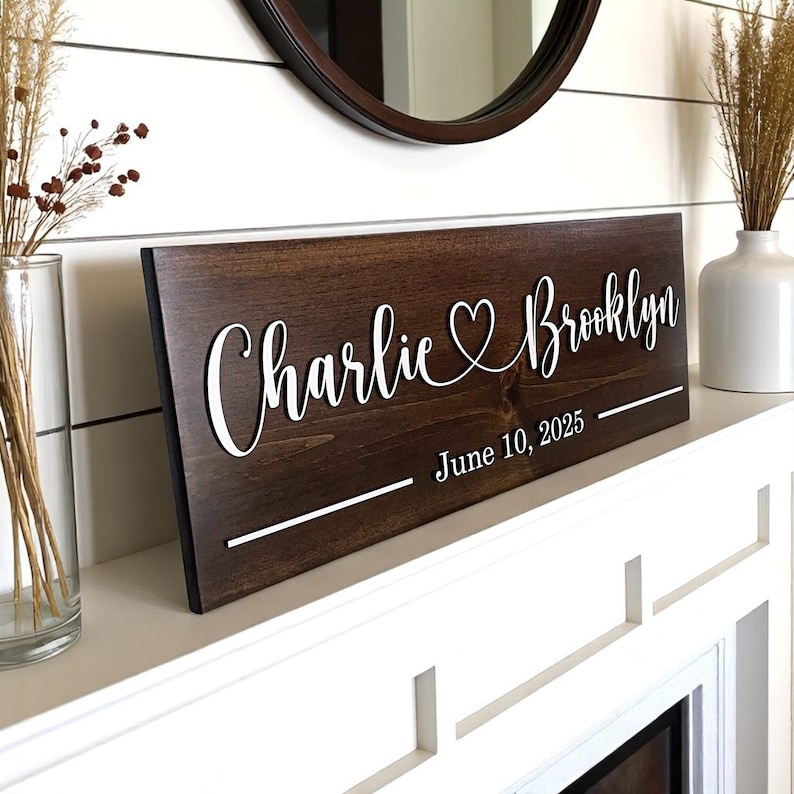 May include: A dark brown wooden sign with white lettering. The sign reads "Charlie & Brooklyn" with a heart symbol between the names, and "June 10, 2025" below. The sign is displayed on a white shelf.