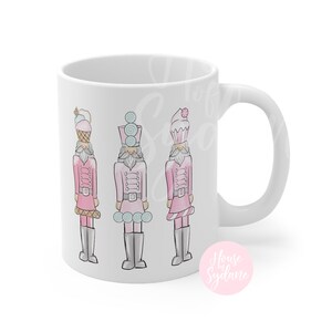 K&ouml;nnte beinhalten: Wei&szlig;e Keramik-Tasse mit einem Design von drei rosa Nussknacker-Soldaten, jeder mit einem Dessert-Themen-Hut. Die Soldaten haben graue Stiefel und G&uuml;rtel. Die Tasse hat einen wei&szlig;en Henkel und die Worte "House by Sydane" in einem rosa Kreis.