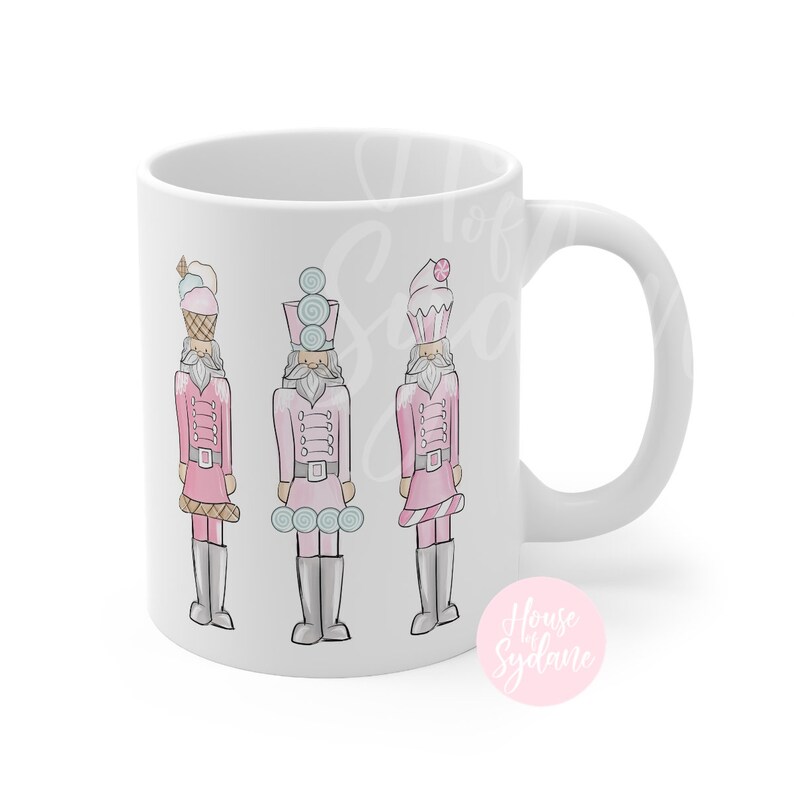 K&ouml;nnte beinhalten: Wei&szlig;e Keramik-Tasse mit einem Design von drei rosa Nussknacker-Soldaten, jeder mit einem Dessert-Themen-Hut. Die Soldaten haben graue Stiefel und G&uuml;rtel. Die Tasse hat einen wei&szlig;en Henkel und die Worte "House by Sydane" in einem rosa Kreis.