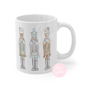 K&ouml;nnte beinhalten: Wei&szlig;e Keramiktasse mit drei Illustrationen von skurrilen Nussknackern. Jeder Nussknacker tr&auml;gt einen Hut und eine Uniform im Dessert-Stil in Pastellfarben. Die Tasse hat einen gebogenen Griff und eine gl&auml;nzende Oberfl&auml;che.