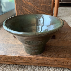 May include: A handmade, green ceramic bowl with a flared rim and a textured, dark brown base. The bowl has a glossy finish and is displayed on a wooden surface. The bowl's design is simple and elegant, suitable for serving or display.
