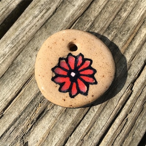May include: A round ceramic pendant with a beige background and a red flower with a black outline. The flower has a white center.