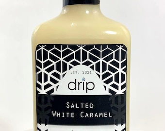 Op de afbeelding: Een doorzichtige glazen fles met een zwarte dop en een wit etiket. Het etiket zegt "drip" en "Salted White Caramel Gourmet Syrup, Creamer, & Dip". De fles bevat een lichtbruine vloeistof en staat op een wit oppervlak.