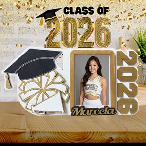 May include: A graduation-themed photo frame featuring the text CLASS OF 2026 in gold glitter. The frame includes a cutout of a graduation cap, a megaphone, and a pom-pom. A rectangular opening displays a photo of a person, with the name Marcela below.