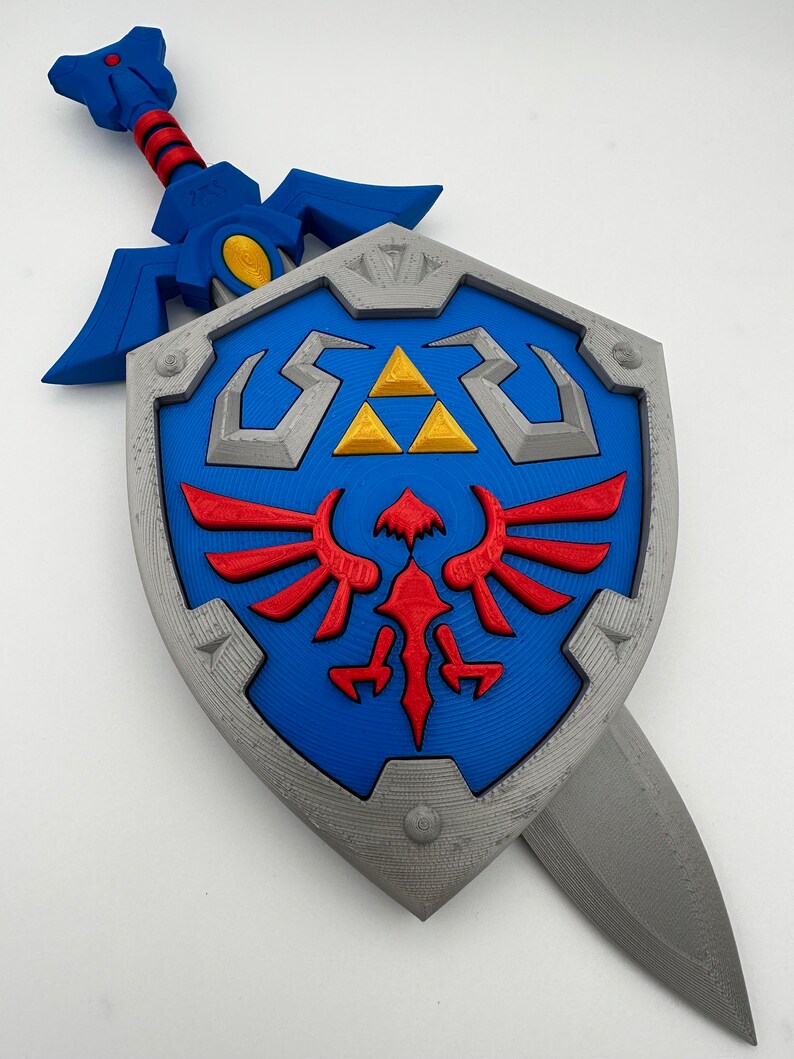May include: A blue and silver plastic replica of the Master Sword and Hylian Shield from the Legend of Zelda video game series. The sword is attached to the shield.