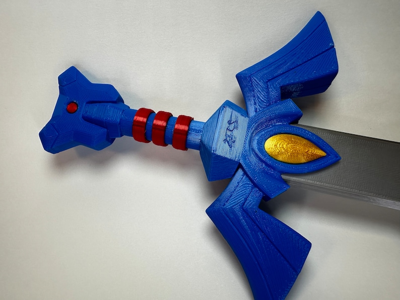 May include: A blue and gold plastic sword with a red and white handle. The sword has a triangular blade with a gold emblem on the front.