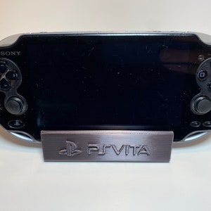Peut inclure: Une console de jeu portable PlayStation Vita noire avec un support gris sur lequel est inscrit "PS VITA".