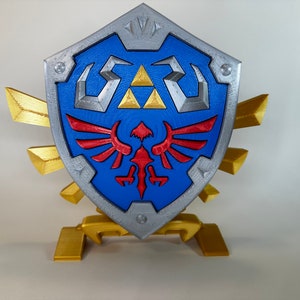 May include: A 3D printed replica of the Hylian Shield from the Legend of Zelda video game series. The shield is blue with a silver trim and features the Triforce symbol and a red and gold winged creature. The shield is mounted on a gold stand.