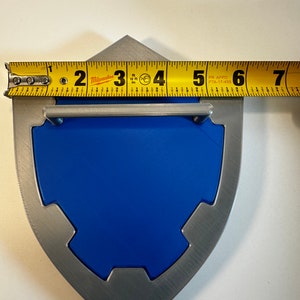May include: A silver and blue shield with a handle. The shield is approximately 6 inches wide. The shield is made of metal and has a textured surface.