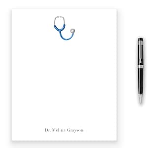 May include: A white notepad with a blue stethoscope illustration and the text "Dr. Melina Grayson". A black pen, gold scissors, and gold binder clips are also visible, along with pink roses.