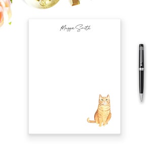 Può includere: Un blocco note bianco con il nome "Maggie Smith" stampato in alto. Un'illustrazione ad acquerello di un gatto tabby arancione si trova in fondo al blocco note.