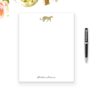 Puede incluir: Un bloc de notas blanco con una ilustración de leopardo dorado y el nombre "Madeline Robinson" impreso en la parte inferior. El bloc de notas está sobre una superficie blanca con un bolígrafo dorado, tijeras y clips.