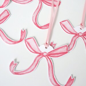 May include: Pink watercolour bow gift tags with a hole punched at the top. The tags are attached to pink ribbon.