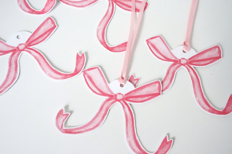 May include: Pink watercolour bow gift tags with a hole punched at the top. The tags are attached to pink ribbon.
