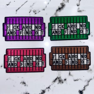 May include: Four rectangular embroidered patches featuring a grid pattern in purple, green, pink, and brown. Each patch displays the white text FUCK AROUND AND FIND OUT arranged in a crossword style across the center, with black borders surrounding each.