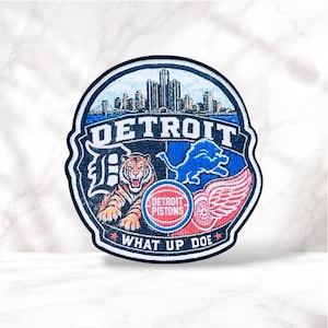 May include: A circular embroidered patch featuring the Detroit skyline, a tiger, a blue lion, a red winged wheel, and a basketball logo with the text Detroit Pistons. The patch includes the text Detroit and What Up Doe in white lettering.