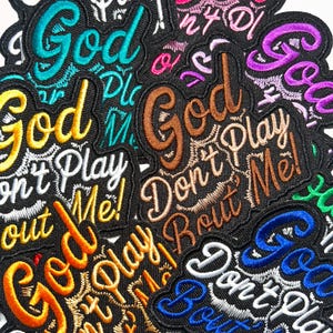 May include: A collection of embroidered fabric patches featuring the text God Don't Play About Me! in various thread colors including teal, gold, white, brown, purple, orange, green, and blue, all set against a black background fabric.