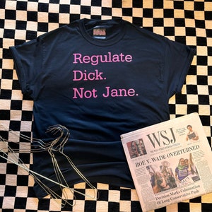 May include: A black t-shirt with the text "Regulate Dick. Not Jane." in pink. A copy of the Wall Street Journal newspaper is next to the shirt. The newspaper headline reads "ROE V. WADE OVERTURNED".