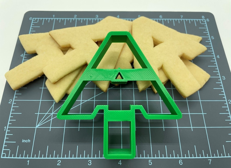 Puede incluir: Un cortador de galletas de &aacute;rbol de Navidad de pl&aacute;stico verde sobre una estera de corte negra con varias galletas de az&uacute;car simples cortadas con el cortador.