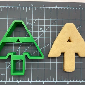 Puede incluir: Un cortador de galletas de pl&aacute;stico verde con forma de &aacute;rbol de Navidad, y una galleta de color marr&oacute;n claro con forma de &aacute;rbol de Navidad.