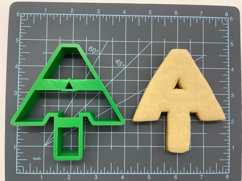 Puede incluir: Un cortador de galletas de pl&aacute;stico verde con forma de &aacute;rbol de Navidad, y una galleta de color marr&oacute;n claro con forma de &aacute;rbol de Navidad.