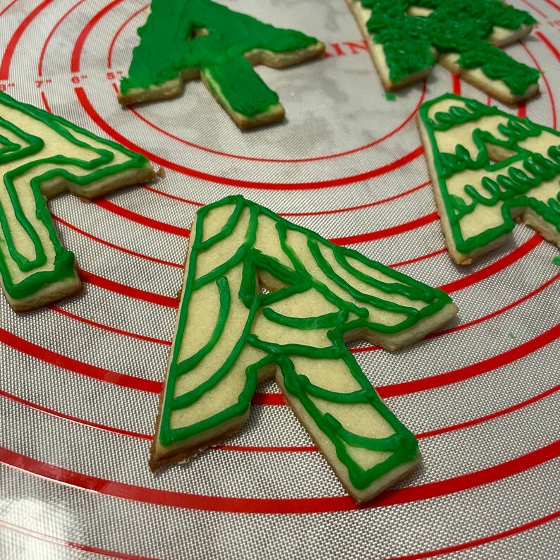 Puede incluir: Cuatro galletas de az&uacute;car glaseadas verdes y blancas con forma de &aacute;rbol de Navidad. Las galletas est&aacute;n sobre una estera de horneado de silicona a rayas rojas y blancas.