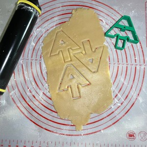Puede incluir: Primer plano de una hoja de masa para galletas con tres cortadores de galletas de &aacute;rbol de Navidad presionados en ella. La masa est&aacute; sobre una estera de hornear de silicona blanca con c&iacute;rculos conc&eacute;ntricos rojos. Un cortador de galletas de &aacute;rbol de Navidad de pl&aacute;stico verde est&aacute; en el lado derecho de la imagen.