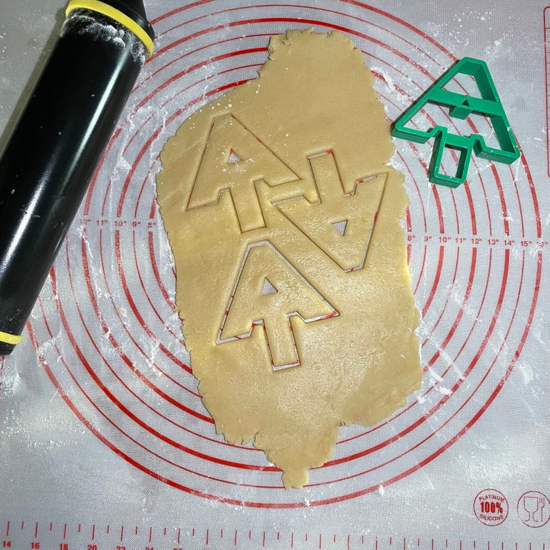 Puede incluir: Primer plano de una hoja de masa para galletas con tres cortadores de galletas de &aacute;rbol de Navidad presionados en ella. La masa est&aacute; sobre una estera de hornear de silicona blanca con c&iacute;rculos conc&eacute;ntricos rojos. Un cortador de galletas de &aacute;rbol de Navidad de pl&aacute;stico verde est&aacute; en el lado derecho de la imagen.