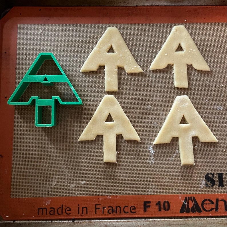 Puede incluir: Cinco cortadores de galletas en una estera de horneado marr&oacute;n. Cuatro tienen forma de &aacute;rbol de Navidad con un agujero en el medio. Uno es un cortador de galletas de &aacute;rbol de Navidad de pl&aacute;stico verde.