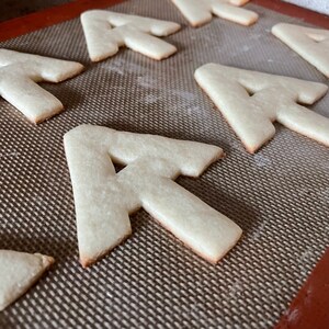 Puede incluir: Primer plano de una bandeja para hornear con varias galletas de az&uacute;car en forma de la letra "A". Las galletas est&aacute;n sin hornear y tienen un color marr&oacute;n claro.