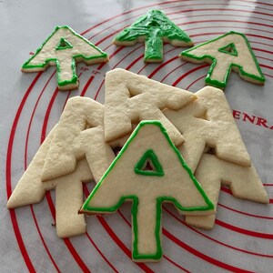 Puede incluir: Una colecci&oacute;n de galletas de az&uacute;car decoradas con glaseado verde en forma de &aacute;rbol de Navidad y la letra A. Las galletas est&aacute;n dispuestas sobre una superficie con un patr&oacute;n rojo y blanco.