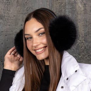 May include: Black fur earmuffs with a black headband. The earmuffs are fluffy and round. The person in the image is wearing a white gilet and a black top.