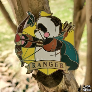 Op de afbeelding: Een gele en gouden emaille pin met een pandabeer die een boog en pijl vasthoudt. De panda draagt een turquoise cape en heeft een gele vogel op zijn hoofd. De pin is gevormd als een geel en gouden schild met het woord "RANGER" op een banner.