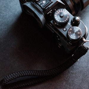 May include: A black camera with a black strap. The camera has a variety of dials and buttons, including a dial for adjusting the aperture and a dial for adjusting the shutter speed.