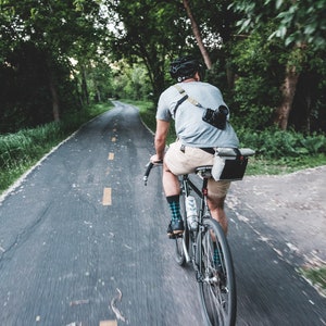 Puede incluir: Una persona monta en bicicleta por un camino pavimentado a través de una zona arbolada. La persona lleva una camisa gris, pantalones cortos color caqui y un casco. La bicicleta tiene un cuadro negro y un sillín de cuero marrón.