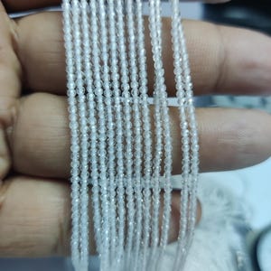 Puede incluir: Múltiples hilos de pequeñas cuentas claras y facetadas. Las cuentas están ensartadas, creando líneas paralelas. Las cuentas parecen estar hechas de un material translúcido, que refleja la luz. Las cuentas se sostienen en una mano.