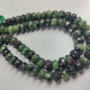 May include: Strands of faceted gemstone beads in shades of green and dark red. The beads are strung together, forming a necklace or bracelet. The gemstones have a polished, faceted surface, reflecting light. The beads are likely used for jewelry making.