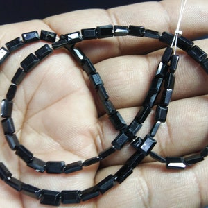 Puede incluir: Un collar de cuentas negras rectangulares. Las cuentas están ensartadas en un hilo blanco fino. Las cuentas son de un color negro oscuro y brillante y parecen pulidas. Las cuentas son uniformes en tamaño y forma, ideales para joyería.