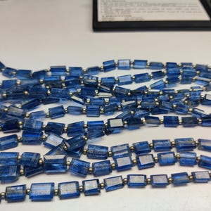 Puede incluir: Hebras de cuentas de piedras preciosas de kianita azul. Cada cuenta rectangular tiene un aspecto pulido y translúcido. Pequeñas cuentas de metal plateado ensartan la kianita. La kianita tiene un color azul profundo y rico.