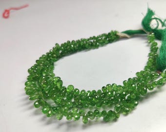 May include: A strand of small, faceted, pear-shaped green gemstone beads arranged in a loose, curved formation. The translucent, vibrant green stones are strung together, with green thread visible at the ends of the bead strand.