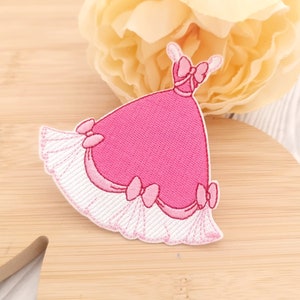 May include: Pink embroidered iron-on patch of a dress with a bow. The dress has a white lace trim and pink bows.