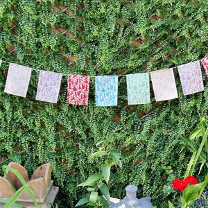 May include: A string of colorful prayer flags with white designs hanging against a green leafy wall. The flags are in shades of blue, green, pink, red, and white.