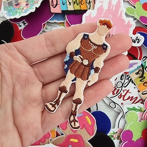 Peut inclure: Une collection d'écussons brodés colorés représentant des personnages et des icônes de Disney. Les écussons incluent une figurine d'Hercule, des châteaux, des silhouettes de Mickey Mouse et du texte qui dit "Disney" et "Always follow your dreams."