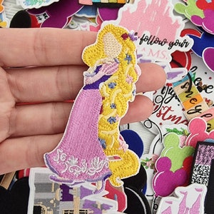 Op de afbeelding: Geborduurde patches met Disney-thema's. Een hand houdt een patch van Rapunzel in een roze jurk vast. Andere patches zijn onder meer een roze kasteel, Mickey Mouse oren en tekst met "Disney", "Believe in Magic" en "Always".