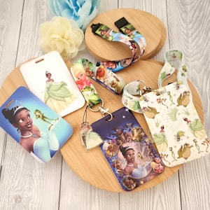 May include: A collection of Disney-themed accessories, including lanyards, keychains, and cardholders. The items feature images of Princess Tiana and other characters in various designs. The accessories are arranged on a wooden surface with decorative flowers.