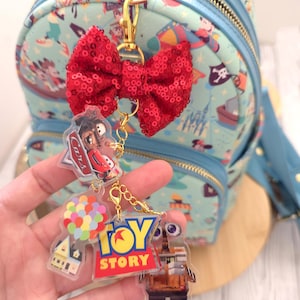 Puede incluir: Un llavero de metal dorado con un lazo rojo de lentejuelas y cuatro dijes de acrílico transparente que representan personajes y logotipos de películas de animación, incluyendo un coche de carreras rojo, una casa con globos y un robot.