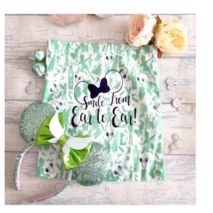May include: A pair of sequined mouse ears with a green bow and a matching green and white patterned fabric. The fabric has a print of silhouettes and the text "Smile From Ear to Ear!"