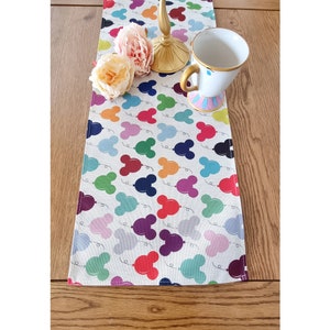 May include: A white table runner with a colorful Mickey Mouse balloon pattern. The runner is decorated with a gold candlestick and a white teacup with a pink and blue floral design.